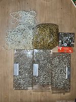 Photo showing an assortment of Crate and Barrel Christmas decorations arranged on a flat surface including white and gold bead strands, three trays of silver jingle bells, and a small box of flocked ornaments