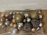 Photo showing an assortment of spherical Christmas ornaments in multiple colors including gold, silver, bronze, and red, some glittered and metallic, along with two woven rattan decorative balls.