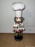 Front view of nutcracker wearing chef’s hat and holding decorated gingerbread house tray