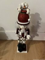 Front view showing the nutcracker with red glittered hat topped by a white snowman figure, holding a small gift with a bow, standing on a white base.