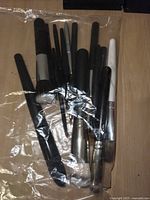Assorted makeup brushes including large, medium, and small brushes with black, silver, and white handles inside plastic bag on wooden surface.