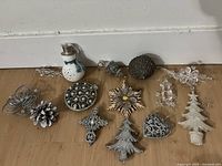 Photo showing the entire collection of 13 items placed on the floor against a white baseboard including plastic snowman, metallic pine cone, glitter snowflake, multiple metal and clear ornaments