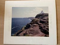 Color photograph mounted on cream mat showing rocky cliff coastline and lighthouse on top under partly cloudy sky.