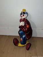 Side view of colorful sitting clown figurine with red jacket, yellow hat, and large shoes, sitting on floor against a white wall.