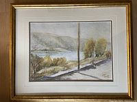 Framed signed watercolour landscape painting showing a river scene with trees and hills in the background.
