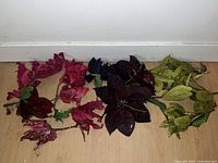 Photo showing brand new silk flowers in red, purple, and green colors arranged on a wooden floor against a white wall.
