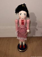 Front view of pink dress women chef nutcracker figurine with black yarn hair and checkered apron standing on a blue and gold base.
