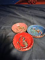 Three small Indigenous Australian bowls with kangaroo designs on red and blue painted ceramic. Displayed on black fabric.