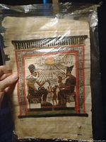 Front view of the Egyptian papyrus painting depicting two seated ancient Egyptian figures with sun rays above and bordered in red and black, held in hand to show details and scale.