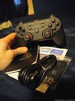 Hand holding the black Gamesir gaming controller with visible buttons and analog sticks. USB charging cable coiled beside the controller with user manuals underneath.