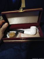 Open red hinged presentation case showing ivory-colored carved meerschaum pipe with black stem and small cork accessory inside.