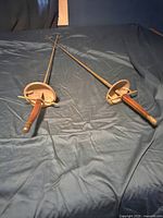 Two fencing swords laid side by side showing wooden handles and metal hand guards, blades are rusty.