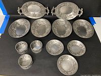 Twelve pewter dishes arranged on a black surface featuring various sizes including two oval trays with handles and round plates with grapevine pattern decoration