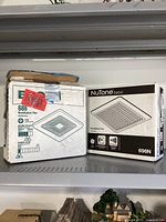 Two bathroom ventilation fan boxes placed on a shelf, one new NuTone Basic 696N box and one opened Broan fan box.
