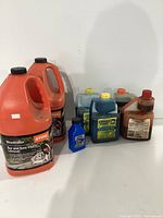 Photo showing front view of 7 oil and lubricant bottles including two large orange Stihl Woodcutter chain lubricant jugs, blue Quaker State, and green Maxima oil bottles.