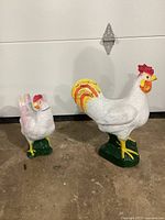Two painted concrete chicken figurines on ground beside a white wall; one taller rooster and one smaller hen with broken neck. Painted white with red, yellow, orange, and green details.