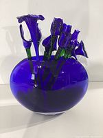 Blue glass flowers and green stems arranged inside a round cobalt blue glass vase. Vase has smooth surface and deep blue color.