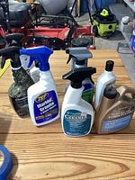 Photo showing six various stone and floor cleaning liquid containers placed on a wooden surface in a garage or workshop setting.