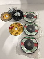 Top view showing seven grinder wheels and a wire brush attachment, various sizes and brands including DeWalt and Hitachi, some sealed in packaging.