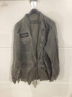 Full view of greenish-gray Harley-Davidson men's jacket hanging showing front button closure, chest pocket with logo patch, and epaulettes on shoulders.