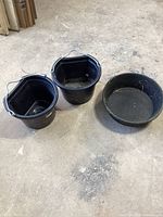 Three black livestock feed buckets on concrete floor showing metal handles and one with a notched rim.