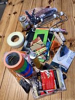 Various painting tools and accessories including roller frames, tape rolls, caulking guns, brushes, sandpaper, and tack cloths organized on a wooden surface.