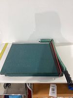 Full view of the heavy-duty green paper cutter table with measurement grid and attached cutting arm with black handle.