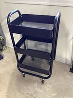 Front left angle view of the black metal rolling storage shelf showing the three mesh shelves and caster wheels.