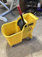 Yellow Rubbermaid mop bucket featuring black wringer with red handle mounted on wheels, viewed from the side with visible wear and dirt marks inside the bucket.