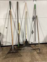 Full view of six yard and garden tools including various rakes, a hoe, and a shovel leaning against a white wall on a concrete floor.