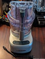 Front view of silver Cuisinart food processor base with attached clear plastic bowl and lid, black control panel with buttons, power cord visible on side.