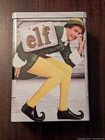 Front side of the rectangular metal tin displaying Buddy the Elf crouching and holding a sign with the movie title 'elf'.