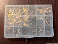 Plastic storage box with various gold-tone jewelry crafting pieces including clasps, rings, and hooks, organized in compartments.