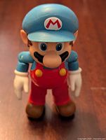 Front angled view of a Super Mario figure with blue cap and red overalls on wooden surface.