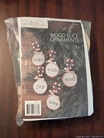 Front package image showing the product name 'Wood Slice Ornaments' with sample decorated wood slices saying 'hope', 'wish', 'noel', 'joy', and 'love' hanging on a green pine background with red and white bead garlands.
