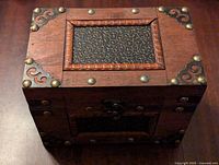 Top angled view of wooden storage box showing framed inset panel with gold swirl pattern and decorative metal corner brackets and rivets.