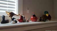A row of various bird figurines on a white surface including wooden, ceramic and plastic ducks and loons, plus a plush bird in the back and small novelties like a letter opener and pencil sharpener shaped like birds.