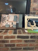 Two canvas bathroom scene paintings leaning against brick hearth alongside Holstein cow figurine