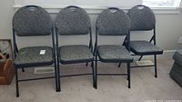 Front view of all four padded folding chairs lined up, showing cloth covered seats and backs and black metal frames.