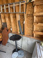 Image showing black plastic bird bath with bird decorative figure, two grey embossed garden stones on the ground, and the green metal shepherds hook in a garage or shed with wooden wall background.
