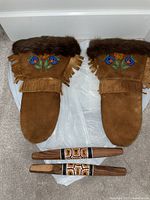 Pair of brown suede mitts with dark fur trim and colorful floral beadwork on the cuffs, next to two wooden cylindrical items decorated with orange and white beadwork.