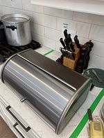 Stainless steel roll-top bread box placed on kitchen countertop near stove and knife block.