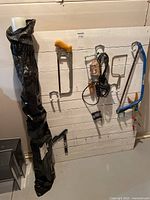 Photo of various household tools hanging on a white wooden slat wall with a black roll of vapor barrier plastic sheeting leaning beside them.