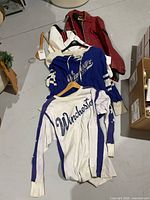 Photo of three 1950s Winchester junior hockey jerseys in white and royal blue, laid out on floor with a red hockey jacket beneath.