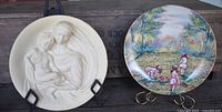 Photo showing both wall plates side by side on stands; left is the white carved Madonna with child, right is colorful floral scene with children picking flowers.