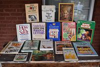 Photo showing 16 assorted books arranged in four rows against a brick wall. Titles include educational, craft, and children's books.