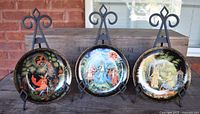 Photo showing the three lacquerware wall plates on decorative iron hangers in front of a wooden box, depicting scenes from Russian fairy tales with vibrant miniature painting on black lacquerware and gold borders.