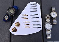 Photo showing 3 wristwatches and various lapel pins including knife designs and Puma logos arranged on white display board and black surface.