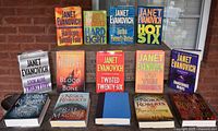 Front view of all 14 hardcover books arranged on a table against a brick wall, showing colorful dust jackets primarily by Janet Evanovich in front, and some Nora Roberts titles alongside.