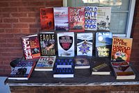 Photo showing 17 military-related books, a mix of hardback and softback, arranged on a table outdoors against a brick wall, covers clearly visible.
